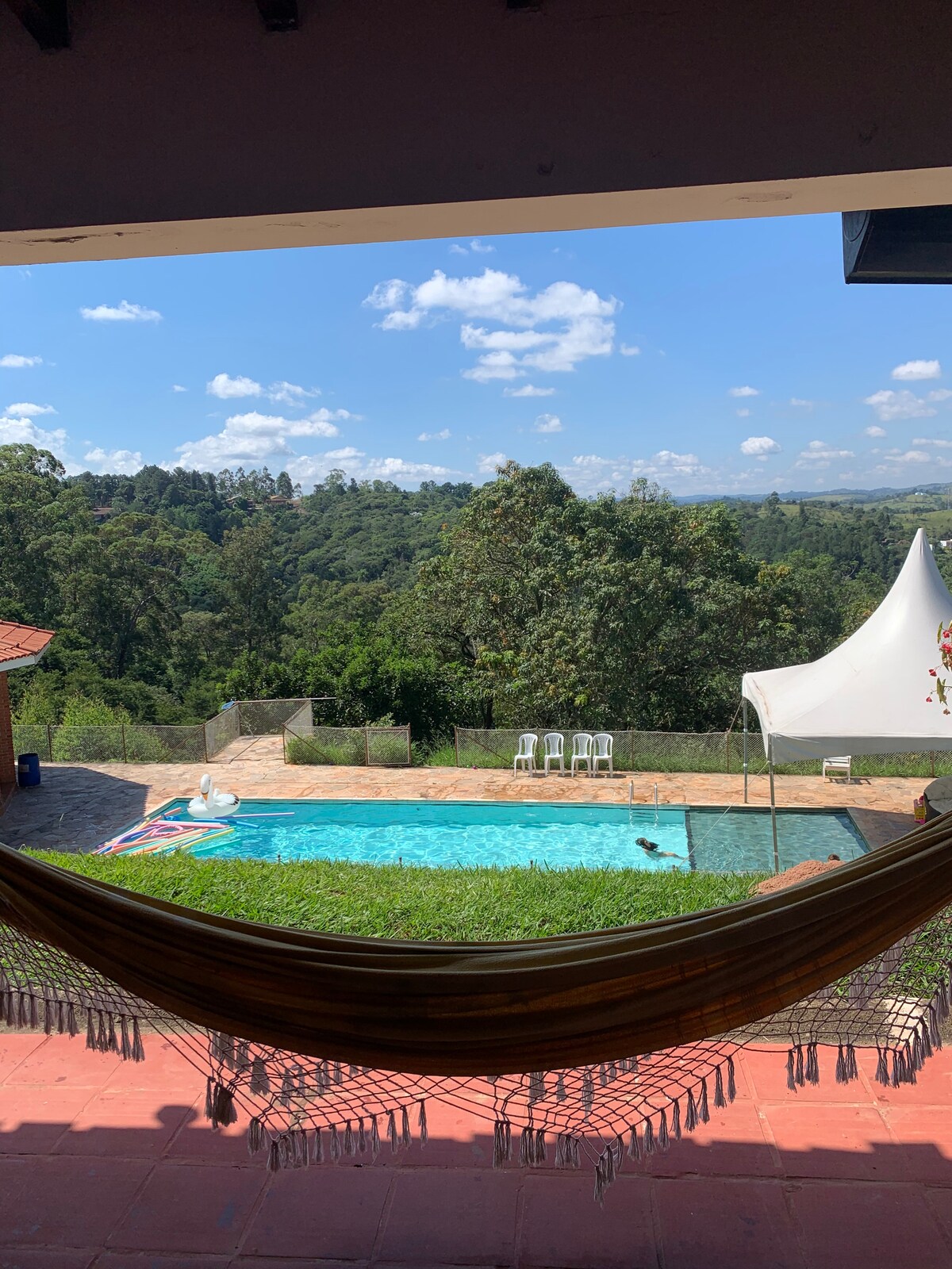 A serene view is presented from a shaded area, showcasing a sparkling pool surrounded by lush greenery and distant hills. A festive tent is visible nearby, along with white lounge chairs situated by the water's edge, creating a welcoming outdoor space.
