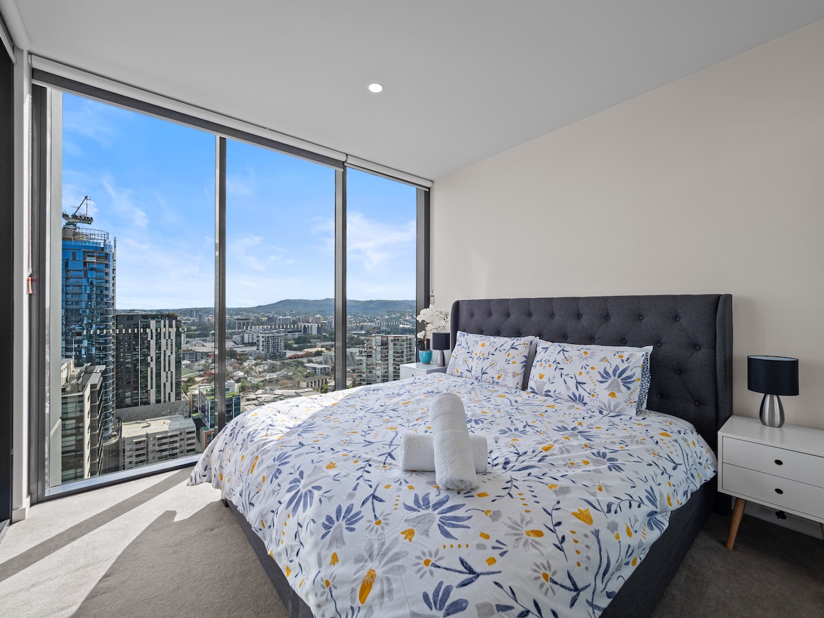 A spacious bedroom features large windows offering expansive views of the cityscape. A king bed is adorned with a floral-patterned duvet and pillows. A small bedside table with a lamp completes the room's layout, which is illuminated by natural light.