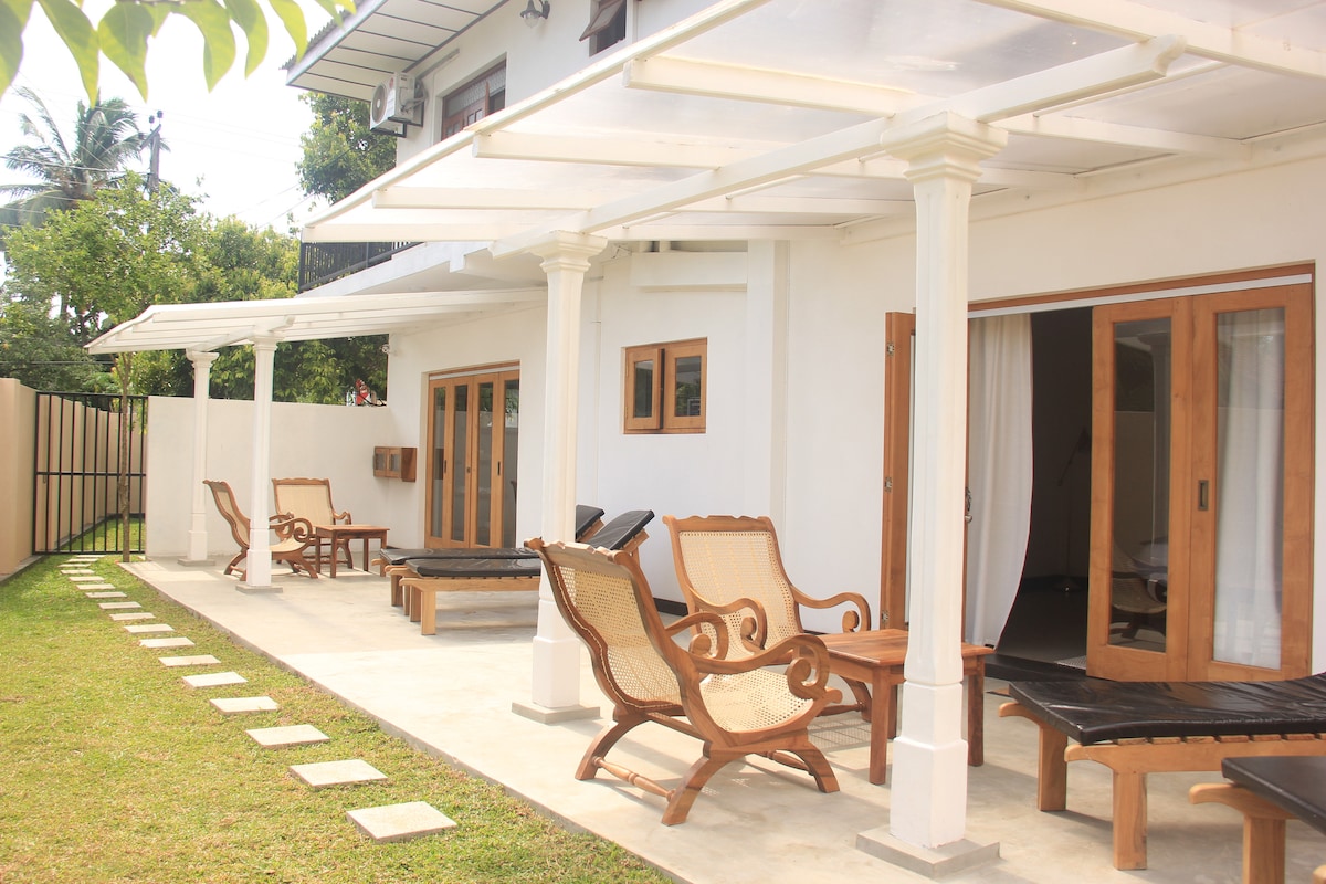 An outdoor seating area is featured, showcasing several lounge chairs alongside a pathway of square stepping stones. A covered patio provides shade, with wooden framed doors on one side leading to an interior space.