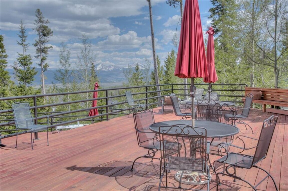 An outdoor deck features a large wooden surface surrounded by trees, offering mountain views in the distance. Several metal chairs are arranged around a circular table, complemented by red umbrellas providing shade. The serene atmosphere invites relaxation in a natural setting.