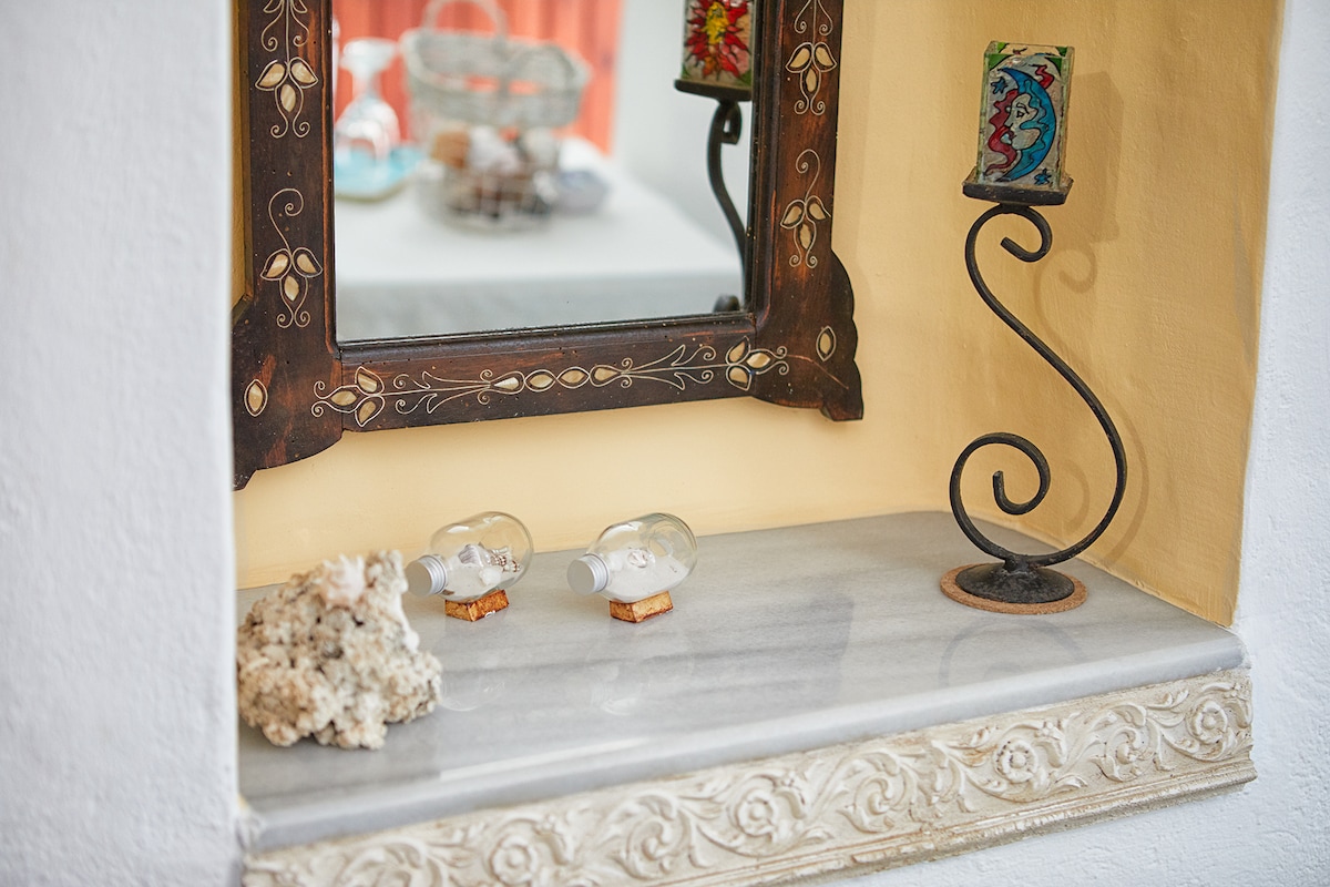 A decorative niche features a polished marble shelf topped with two small glass bottles resting on cork bases. An ornate candle holder stands beside them, while a carved wooden mirror enhances the space, reflecting soft light from the room.