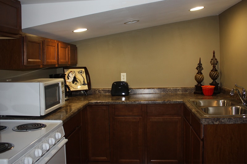 A small kitchen area is equipped with wooden cabinetry and a granite countertop. Appliances include a microwave, toaster, and a stove. A double sink is positioned beneath a wall-mounted light, and decorative items enhance the workspace.