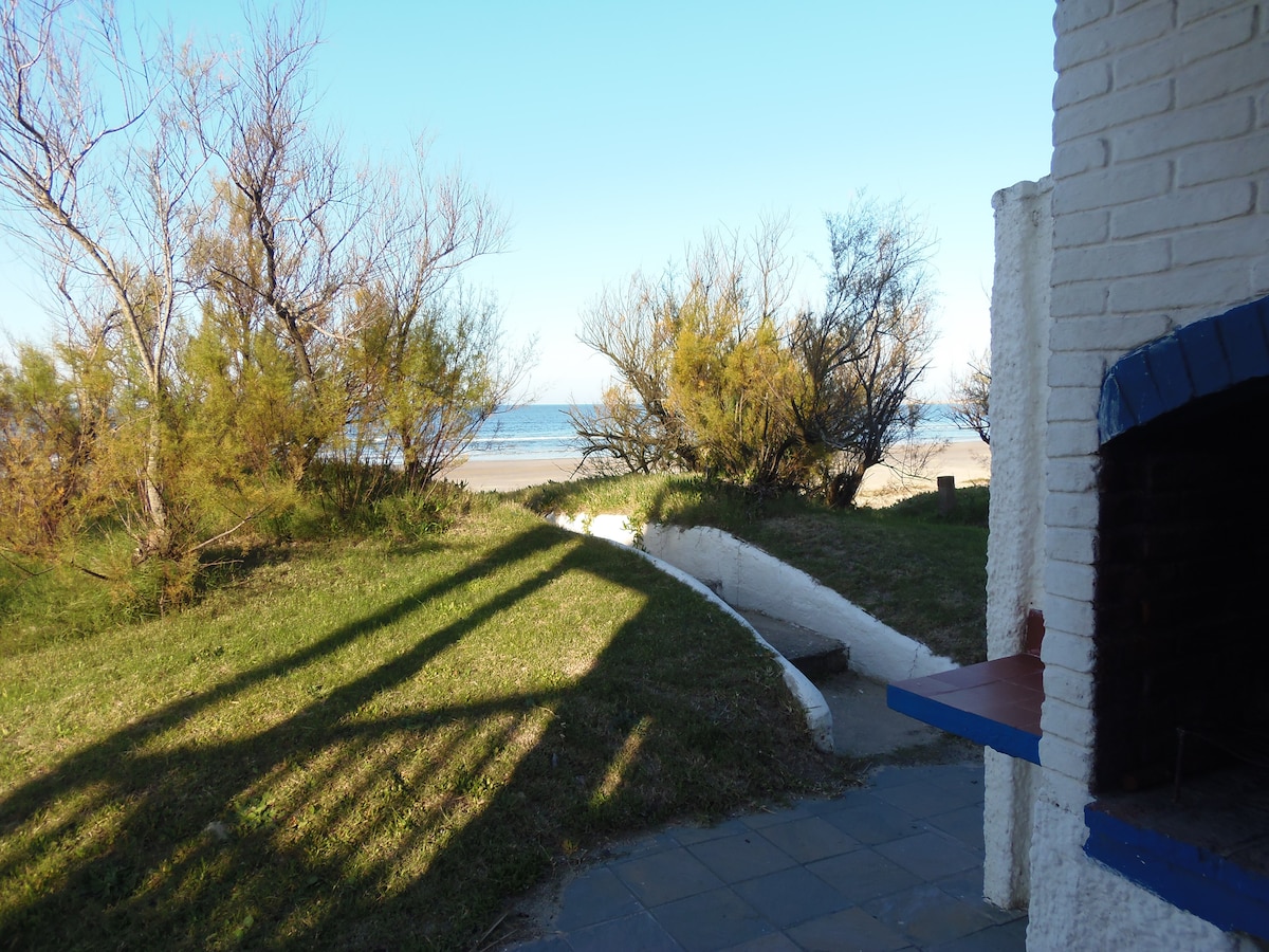 A view is provided of the outdoor area, featuring lush greenery and a stone BBQ. The horizon showcases the ocean, with gentle waves visible. Shadows are cast by nearby trees, enhancing the serene beach atmosphere.
