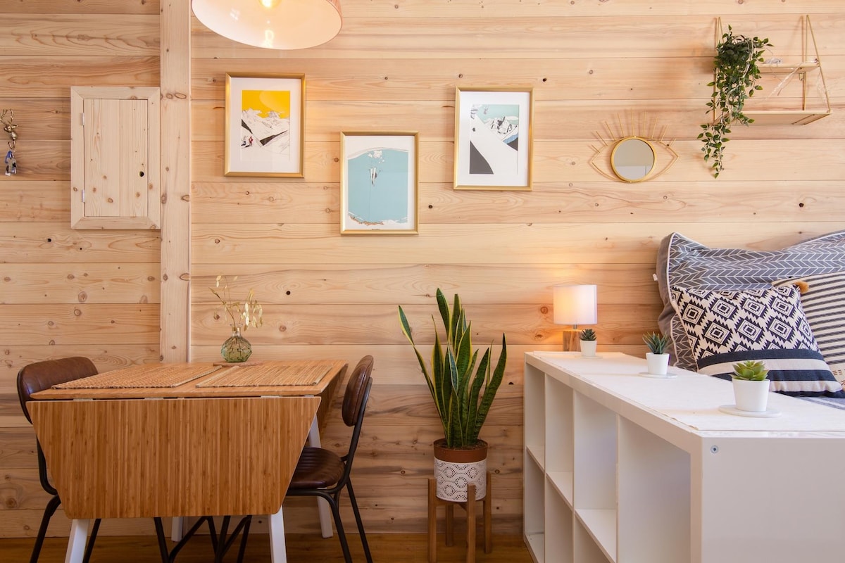 A warm wooden interior displays a dining area featuring a compact table surrounded by four chairs. Wall art in colorful frames adds visual interest, complemented by green plants. Soft lighting from a pendant lamp enhances the cozy atmosphere, and a mirror reflects natural light.
