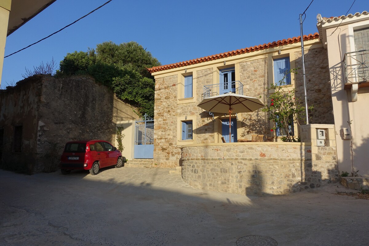 A renovated stone house is shown in a quiet side street. The light-coloured facade features blue shutters and a small terrace with an umbrella. A red car is parked nearby, alongside greenery that enhances the charming setting.