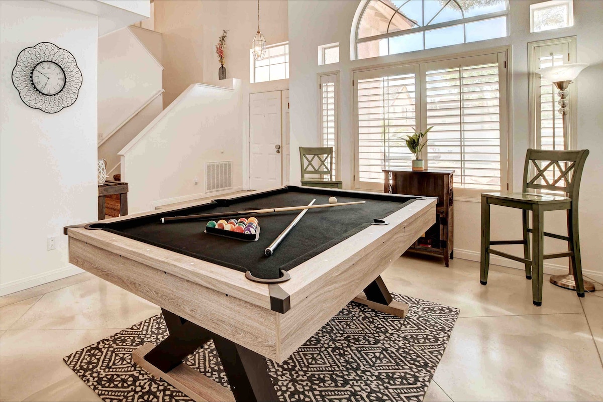 A pool table is positioned in a brightly lit area featuring large windows with plantation shutters. The surrounding space includes a decorative rug and two chairs at a small table, creating an inviting recreational setting.
