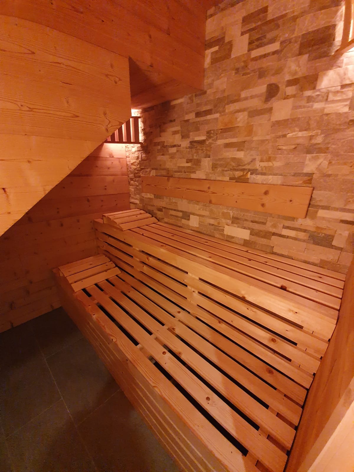 A warm sauna is depicted with wooden slatted benches and stone walls. Soft lighting creates a serene ambiance, enhancing the inviting feel of the space. The wooden construction suggests a cozy, natural atmosphere, perfect for relaxation.