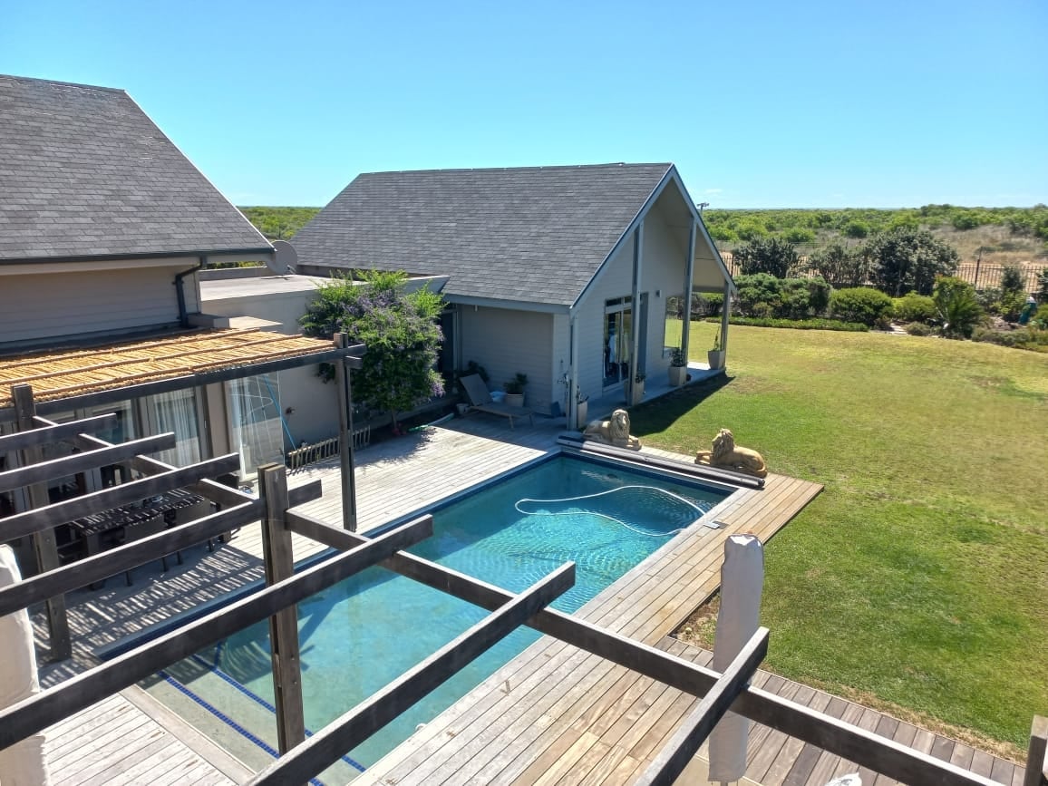 An aerial view of the property showcases a private pool surrounded by a wooden deck. The home features an outdoor area with seating, and lush greenery is visible in the background. The clear blue sky adds brightness to the scene.