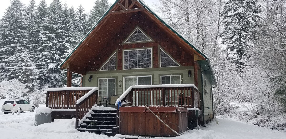 The exterior of a chalet-style cabin is visible, surrounded by a snowy landscape and evergreen trees. A large covered porch with wooden railings leads to multiple glass windows showcasing the interior. A hot tub is positioned to the side, inviting relaxation in winter weather.