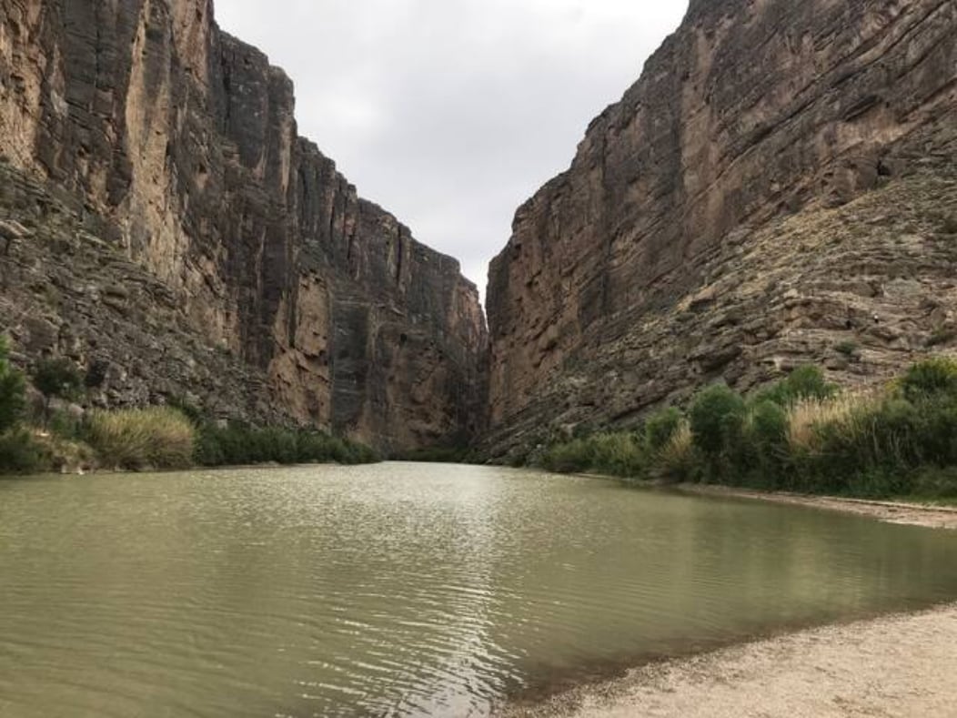 A serene river flows between towering rock formations, with steep, rugged cliffs rising on either side. The water appears still, reflecting the muted sky above. Lush vegetation lines the riverbank, enhancing the natural beauty of the landscape.