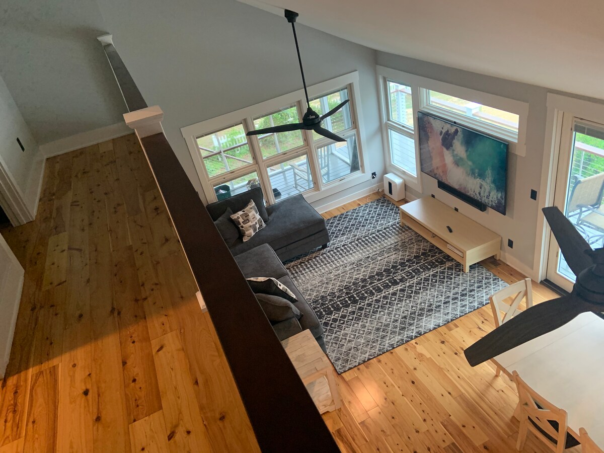 A spacious living area is shown with large windows allowing ample natural light. A comfortable grey sectional sofa faces a light wooden coffee table. A decorative area rug lies beneath, and a view of the outdoor deck is visible through the windows.