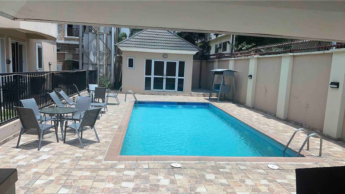 A swimming pool is set within a paved outdoor area, surrounded by a black fence. Lounge chairs and a table with four chairs are arranged beside the pool. A covered seating area is visible, along with an adjoining building in the background.
