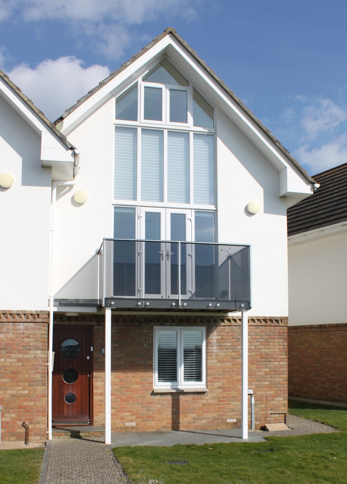 The exterior of the modern home showcases a combination of white walls and brick detailing. A glass balcony extends from the upper level, providing an unobstructed view. The front door features a circular window, with well-kept grass leading up to the entrance.