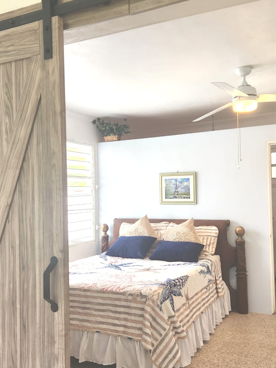 A cozy bedroom presents a wooden bed adorned with a striped blanket and decorative pillows. A ceiling fan hangs overhead, while light filters through jalousie windows. A small plant and a wall art piece enhance the serene environment.