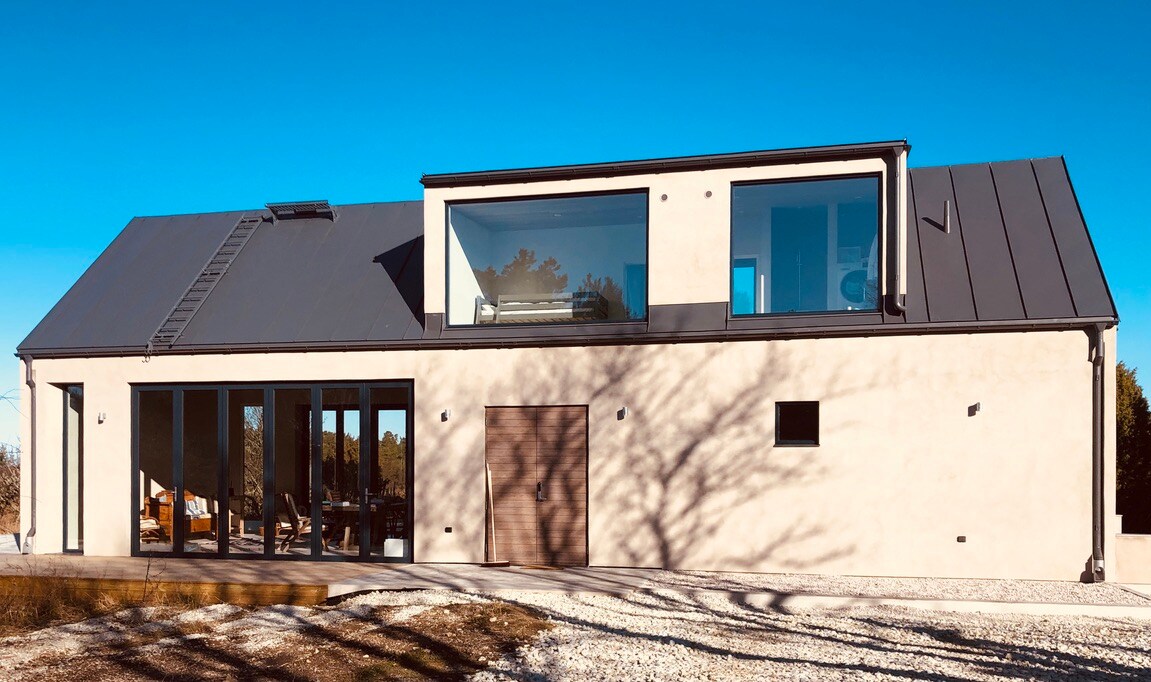 The exterior of a modern two-story villa is presented, featuring large glass windows and a clean design. A mix of light-colored walls and a dark roofing contrasts appealingly. A pathway leads up to the entrance, surrounded by a spacious lawn, framed by the surrounding nature.