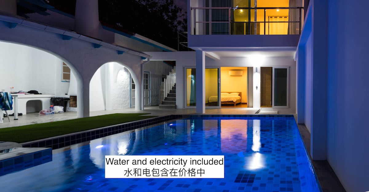 A private swimming pool reflects soft lighting from the surrounding areas. The bungalow's exterior displays a mix of modern architecture, with large windows and a grass lawn. A text overlay indicates that water and electricity are included in the stay.