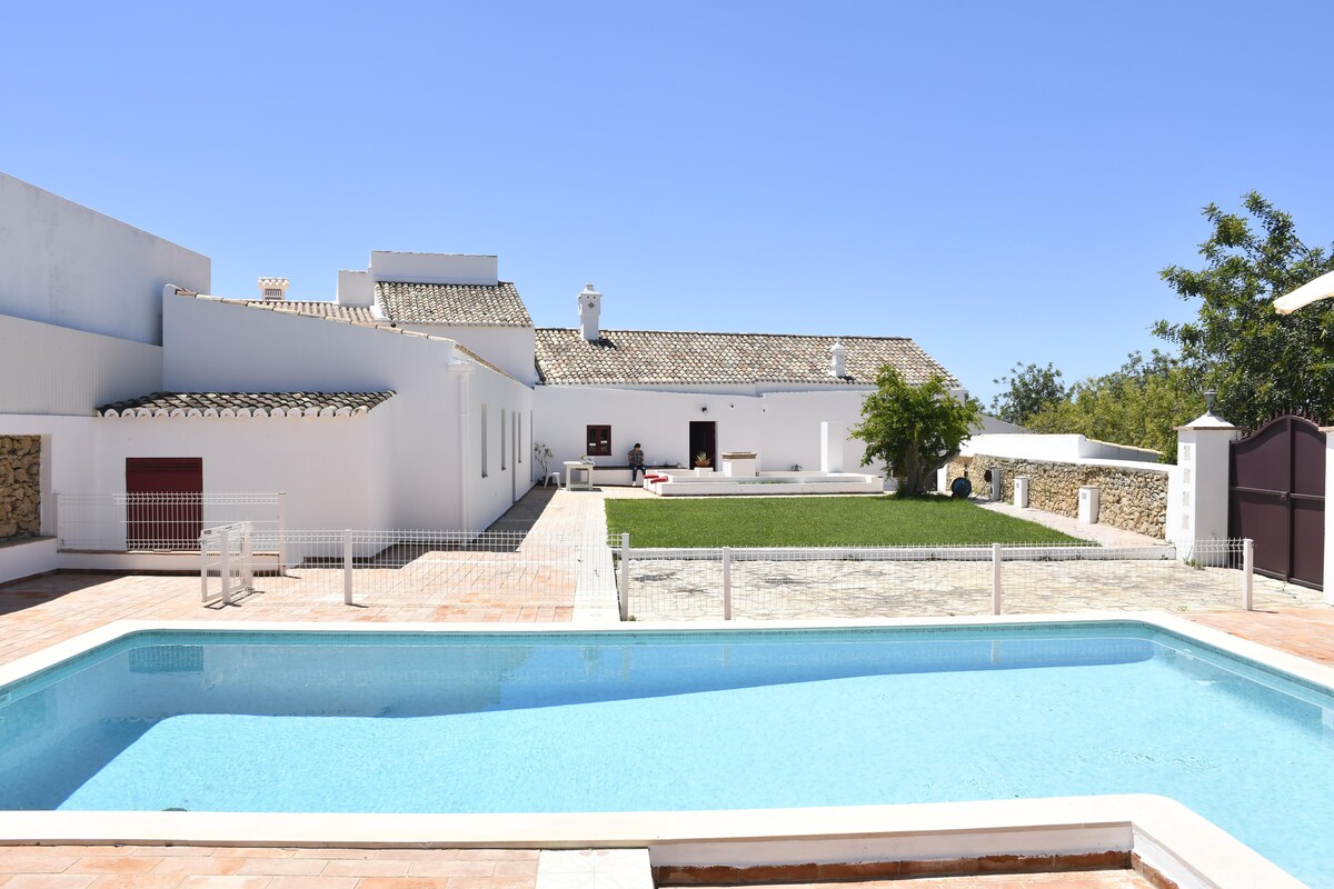 A serene outdoor area features a refreshing swimming pool surrounded by a patio. The spacious lawn offers greenery, with a charming white building in the background that showcases a traditional roof. Clear blue skies enhance the peaceful atmosphere.