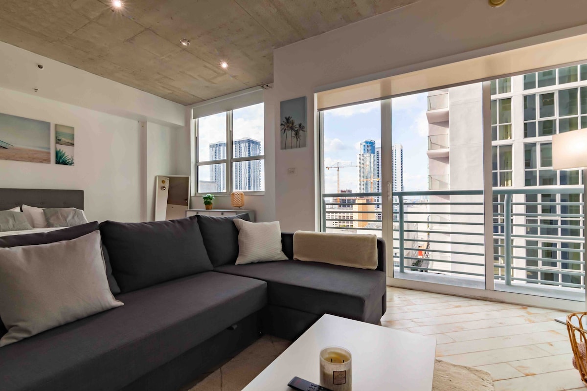 A spacious living area features a dark gray sectional couch adorned with soft pillows. Large windows provide ample natural light and offer views of the city skyline. A light-colored coffee table sits in front of the couch, contributing to the inviting ambiance.