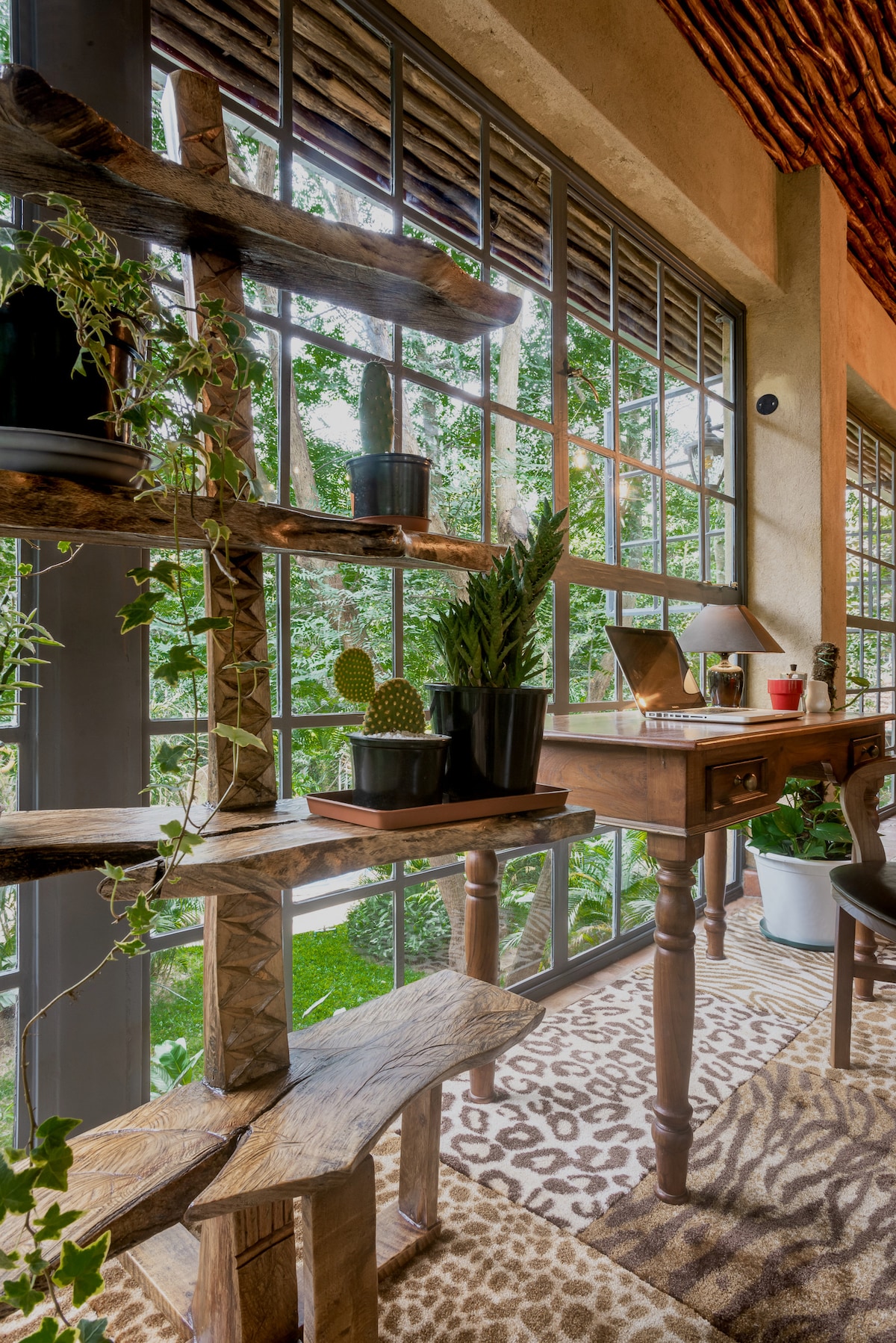 A bright workspace is featured, showcasing a rustic wooden desk beside large windows that allow natural light to flood in. Green plants are displayed on a unique wooden shelf, bringing elements of nature indoors. The room is decorated with a patterned rug, enhancing its inviting atmosphere.