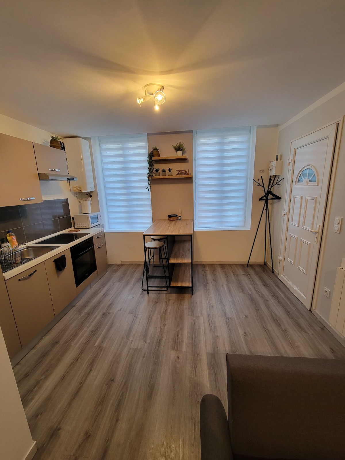 A modern kitchen area is presented with light-colored cabinetry and a countertop. Two barstools are positioned at a small table, and a microwave and refrigerator are visible. The room is filled with natural light from two large windows, complemented by a warm light fixture.