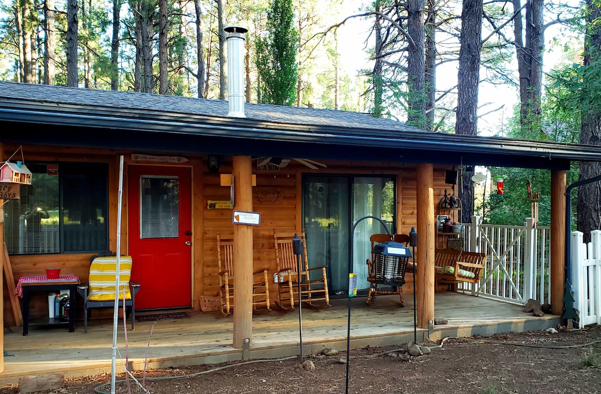 The cabin's exterior features a welcoming red door and a spacious porch adorned with multiple rocking chairs. Pine trees surround the structure, providing a natural backdrop. A chimney is positioned on the roof, indicating a cozy ambiance. The railing adds a touch of charm to the outdoor area.
