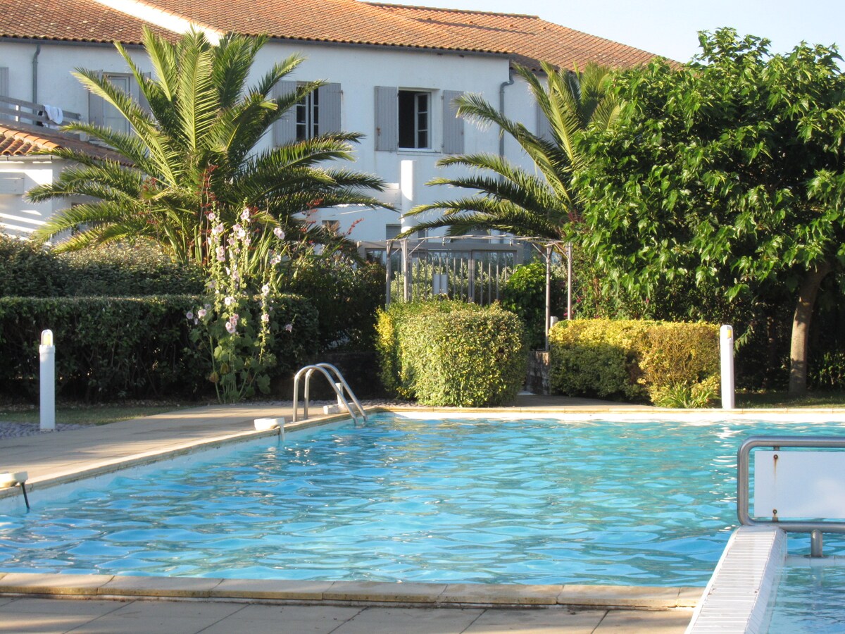 An outdoor swimming pool is surrounded by lush gardens and palm trees. The clear blue water reflects sunlight, enhancing the inviting atmosphere. The pool area features tiled flooring, and various green shrubs and plants create a serene backdrop.