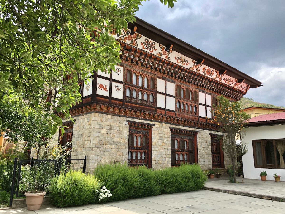 A traditional two-story Bhutanese house is set against a moody sky, featuring ornate wooden balconies and intricate carvings along its exterior. Lush greenery and flowering plants surround the structure, with a paved courtyard visible in front, providing a welcoming natural atmosphere.