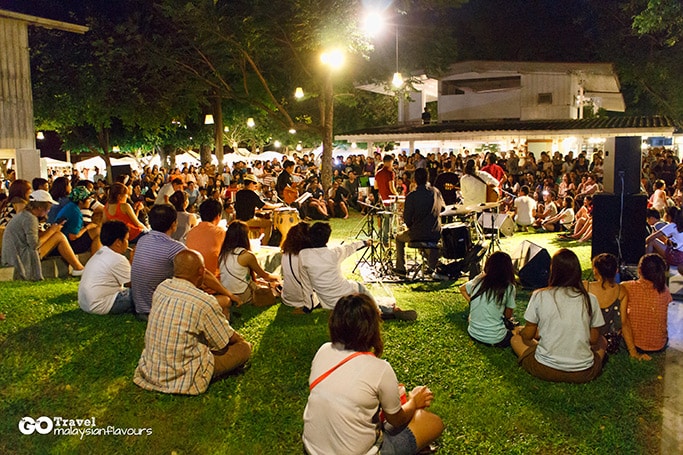 An outdoor gathering takes place in a vibrant green space, filled with engaged attendees seated on the grass. A live performance is being held on a small stage, surrounded by trees and softly lit by hanging lights, creating a lively yet relaxed atmosphere.