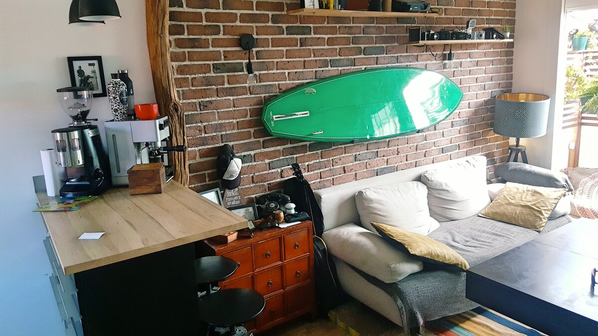 A living area features a modern sofa adorned with cushions, positioned next to a wooden coffee table. A wall with exposed brick showcases a green surfboard, while a compact bar area with stools offers additional seating. A lamp and decorative items complete the space.