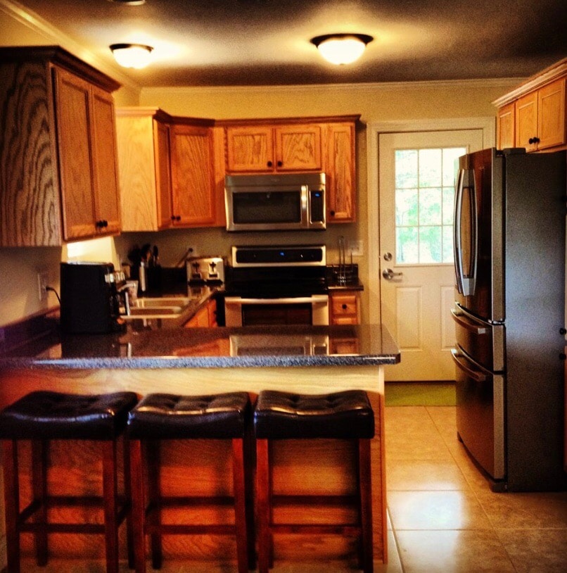 The kitchen features wooden cabinetry and modern appliances, including a microwave and oven. A dark granite countertop extends over the kitchen island, accompanied by three stools. Natural light enters through the door with glass panels, enhancing the functional layout.