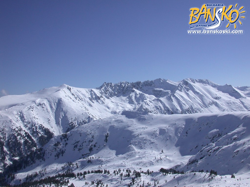 A breathtaking winter landscape showcases vast, snow-covered mountains under a clear blue sky. The terrain features rolling hills and distant peaks, offering a serene view of nature's beauty in a tranquil setting.