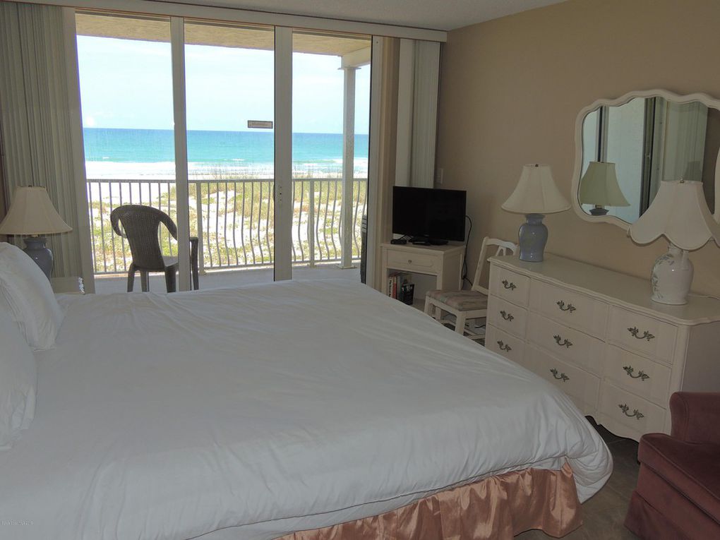 A spacious bedroom features a large bed with crisp white linens. A mirror and television are placed across from the bed, while a chair provides additional seating. Floor-to-ceiling glass doors open to a balcony with ocean views, enhancing the light and airy ambiance.