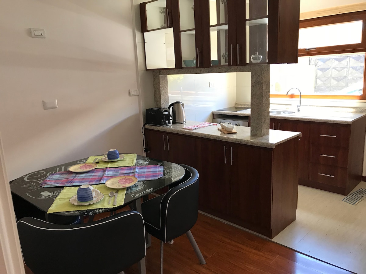 A modern kitchen area features dark wood cabinetry and a granite countertop. A circular dining table is set for two, with vibrant place settings and cups. The kitchen is equipped with an electric kettle and has large windows allowing natural light.