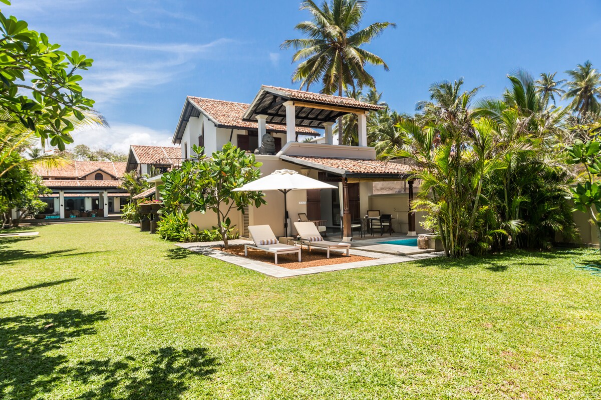 A spacious garden surrounds the villa, featuring well-maintained grass and tropical foliage. Comfortable sun loungers are positioned by the pool, under the shade of a large umbrella. The two-story villa, with a tiled roof, complements the serene outdoor setting.