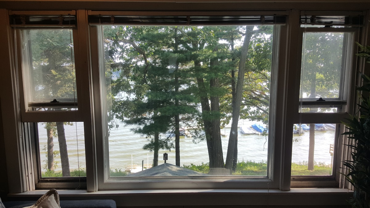 Large windows provide a view of Petite Lake, framed by tall trees. Boats are visible on the water, and gentle waves can be seen lapping against the shoreline. Natural light fills the space, enhancing the serene lakeside atmosphere.