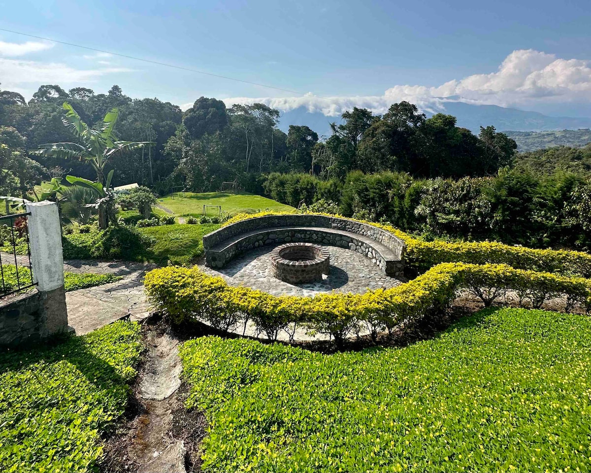 A circular stone fire pit is surrounded by neatly trimmed greenery, providing a cozy gathering area. Lush gardens and trees are visible in the background, with rolling hills and a clear sky creating a serene rural landscape.