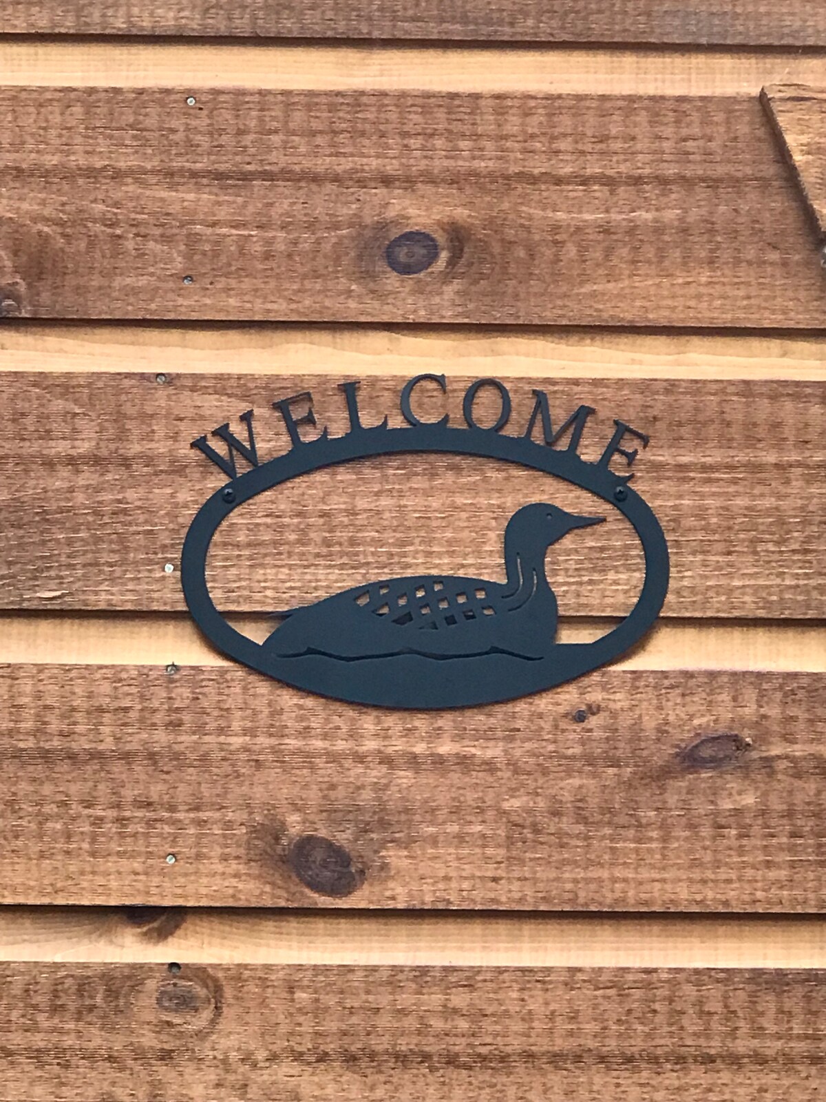 A welcome sign crafted from metal is mounted on a wooden exterior. The sign features a silhouette of a loon, framed within an oval shape that complements the natural texture of the wood.
