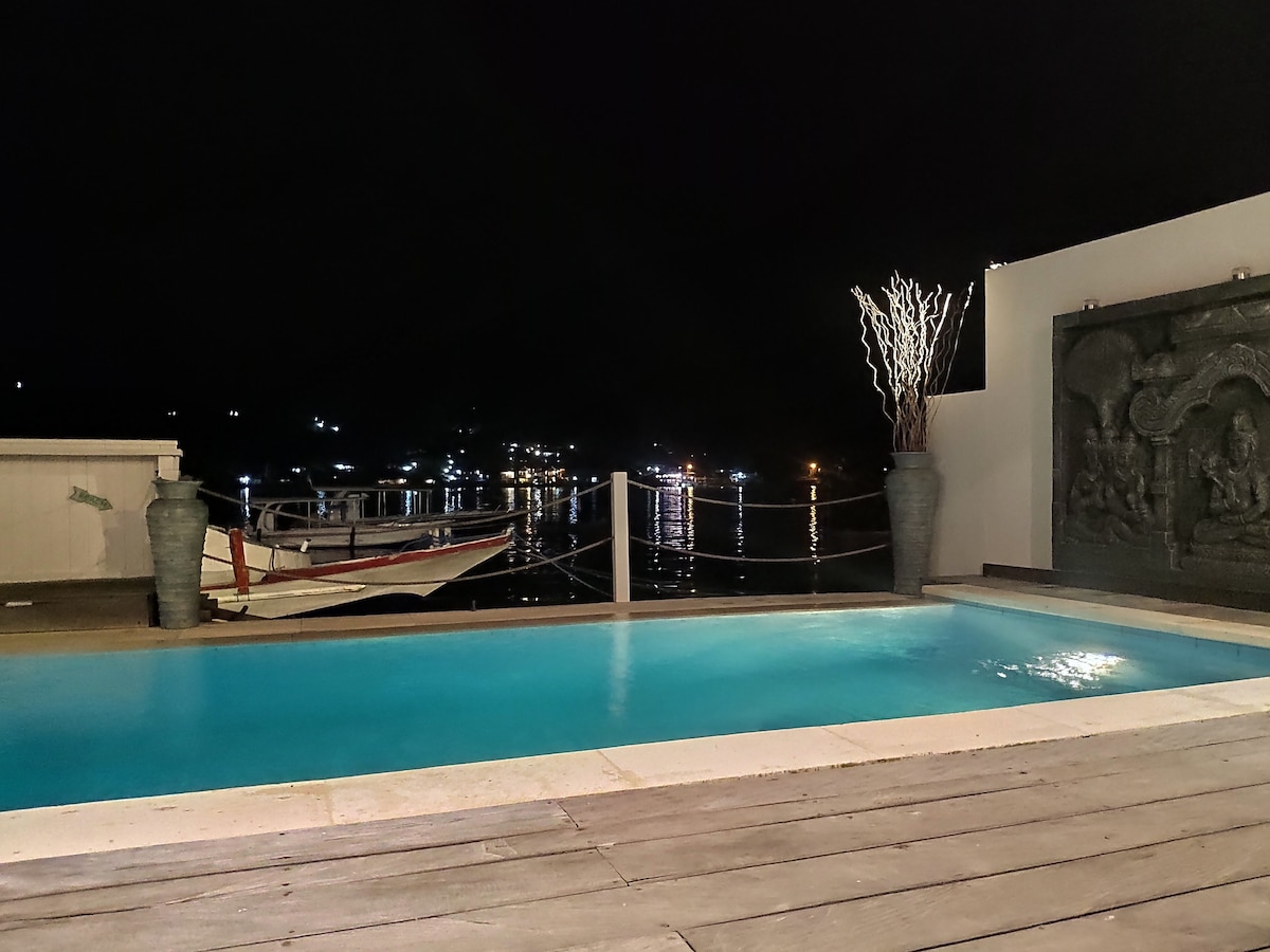 The serene pool area is illuminated softly at night, reflecting the surrounding water and distant lights. Elegant planters with tall, bare branches are positioned alongside the pool, while a traditional boat is gently anchored nearby, enhancing the tranquil atmosphere.