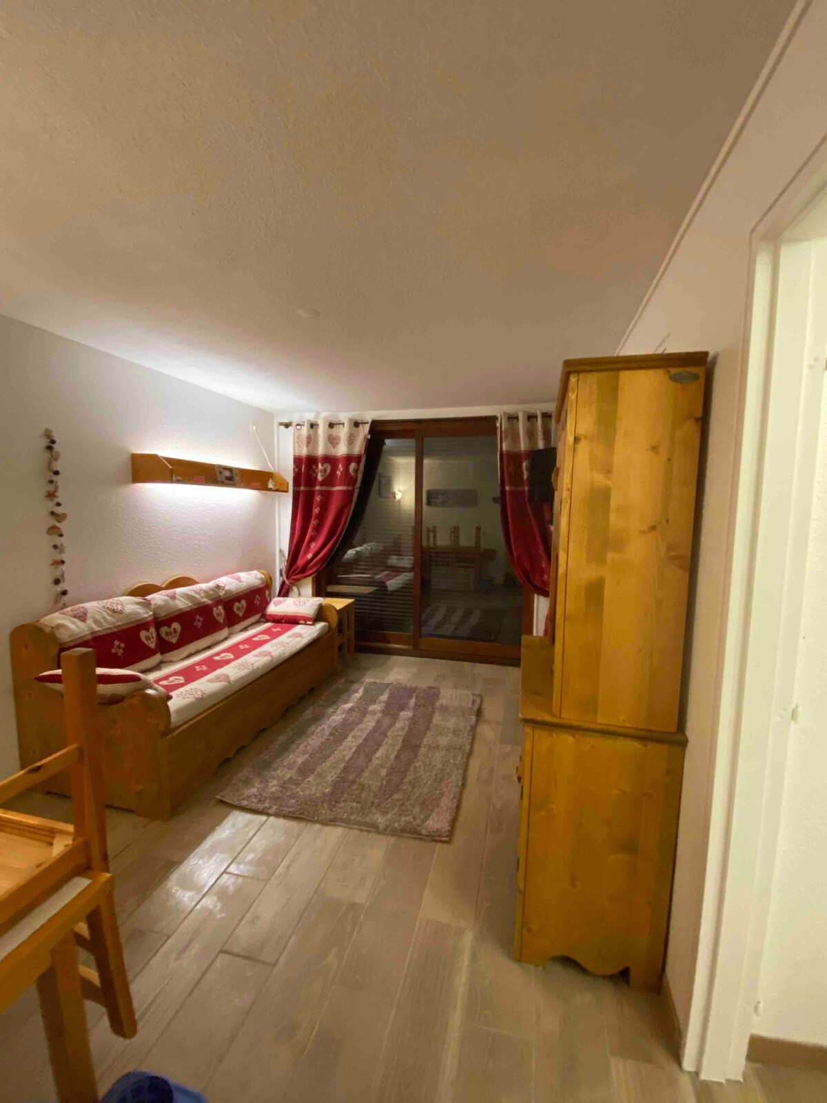 A cozy living space features wooden furnishings, including a seating area with decorative cushions. A rug adds warmth to the floor, while large glass doors lead to a balcony, allowing natural light to filter into the room. Curtains frame the doors, enhancing privacy.
