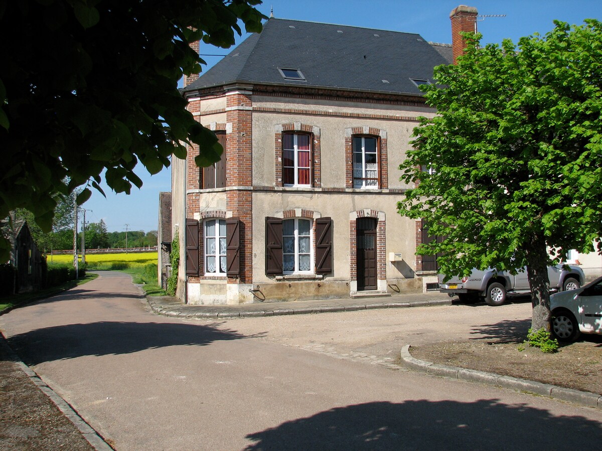 A charming three-story house is seen from the corner of a quiet street, showcasing a classic facade with brick detailing and shuttered windows. Lush greenery surrounds the building, with unobtrusive parked cars visible along the side, and fields of vibrant yellow flowers in the distance.