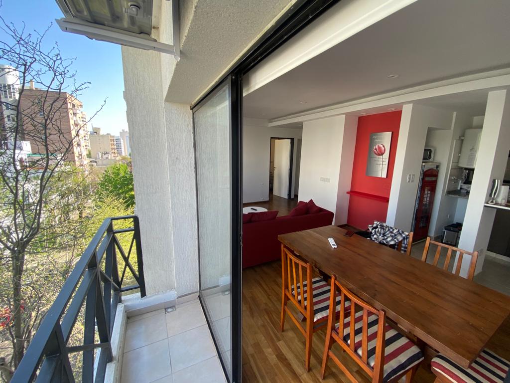 A balcony view reveals a dining table with wooden chairs alongside a large window opening to the interior living space. The bright and airy environment is enhanced by natural light, showcasing a cozy seating area in the distance and a well-equipped kitchen beyond.