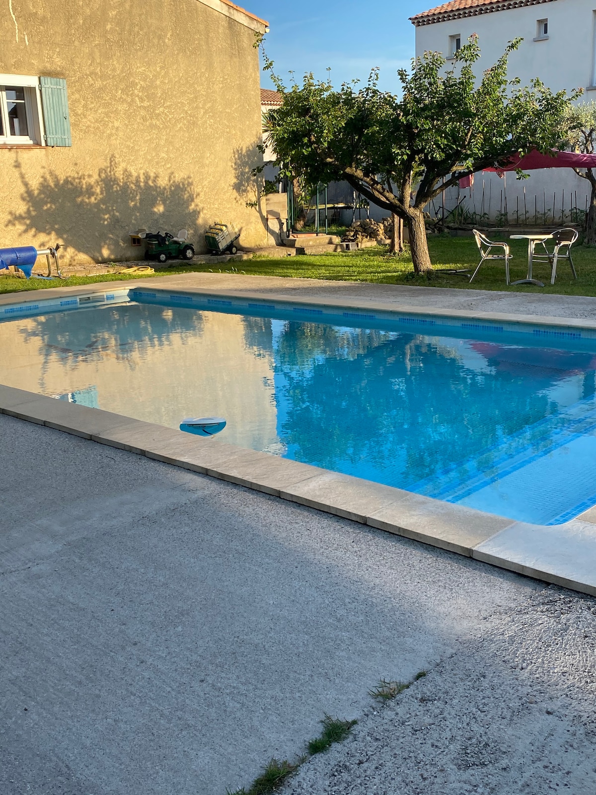A refreshing swimming pool is set within a well-maintained garden, surrounded by greenery. Sunlight reflects off the water's surface, while a few lounge chairs are positioned nearby. The area is designed for relaxation, with trees providing shade and a calm atmosphere.