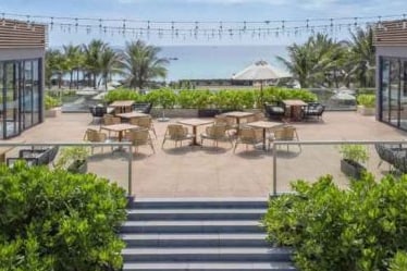 An outdoor terrace features several seating arrangements with tables and chairs surrounded by lush green plants. A large umbrella provides shade in one area, while the ocean is visible in the background under a clear blue sky.