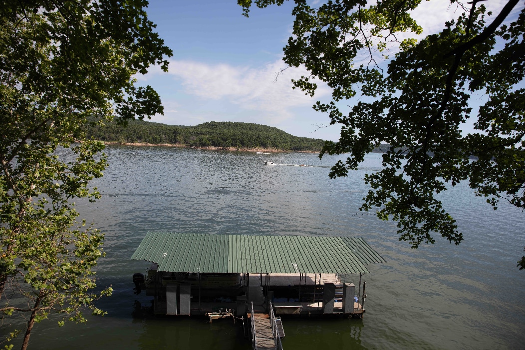 A spacious dock extends over the calm waters of Lake Tenkiller, surrounded by lush greenery. The dock features a metal roof and provides access to the water, with distant hills visible on the horizon under a clear sky.
