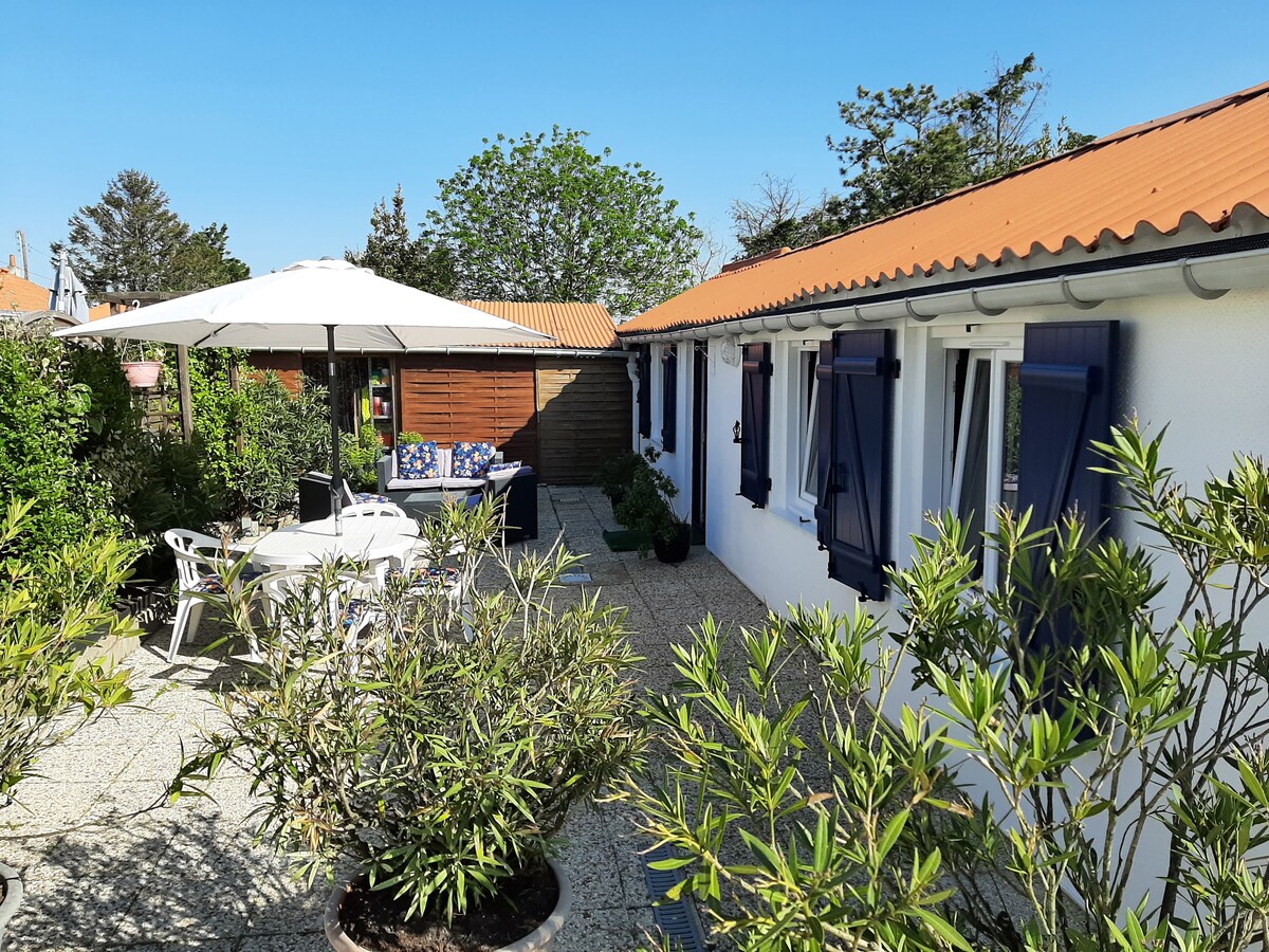 A charming exterior space is filled with greenery, featuring a patio with a large umbrella and dining table. The white house, accented by blue shutters, is surrounded by well-maintained plants, contributing to a welcoming atmosphere. A cozy seating area can be seen in the background.