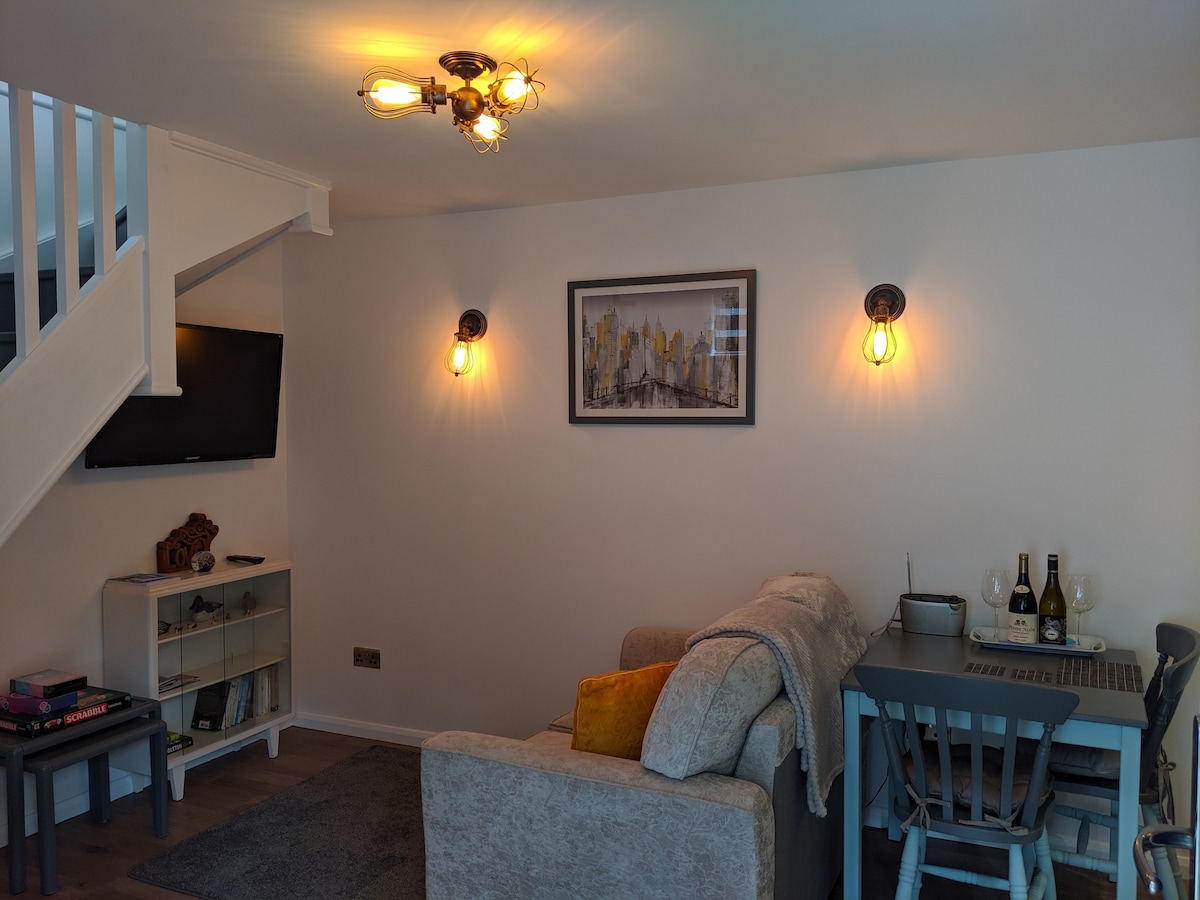 A cozy living and dining area features a comfortable couch adorned with a soft throw. A dining table for two sits nearby, complemented by a bottle of champagne and glasses. Ambient lighting creates a welcoming atmosphere, with a wall-mounted TV providing entertainment options.