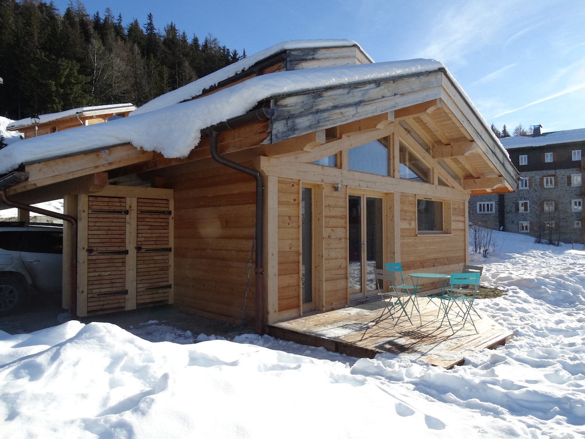 A wooden chalet is displayed with a sloping roof, complemented by a small terrace featuring two chairs. Snow covers the ground, and trees are visible in the background. The structure includes large windows, inviting natural light into the interior space.