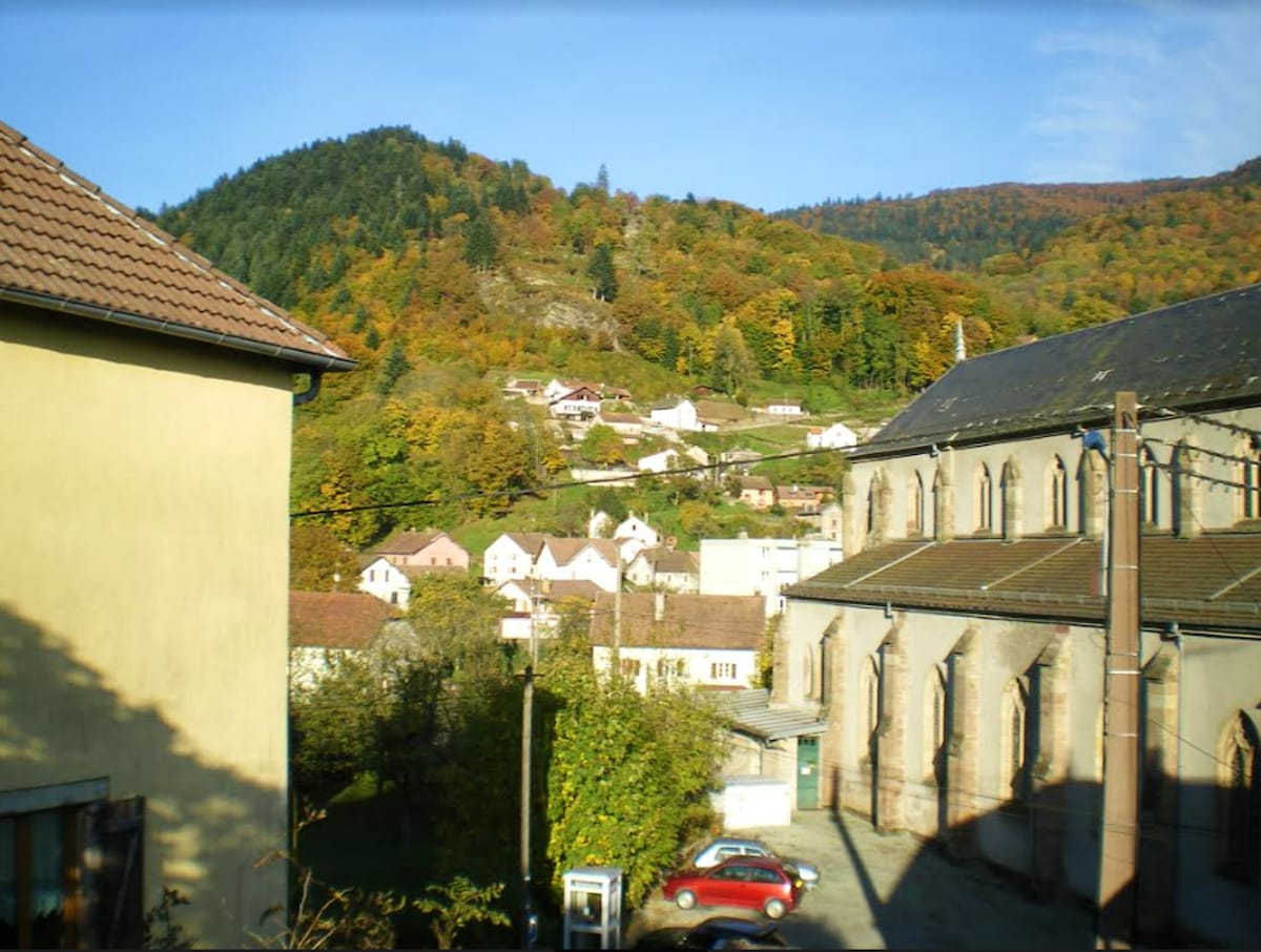 A scenic view captures the vibrant foliage of nearby hills, with trees displaying autumn colors. Charming hillside houses are visible, nestled among the landscape. A prominent building with arched windows stands in the foreground, framed by a clear blue sky.