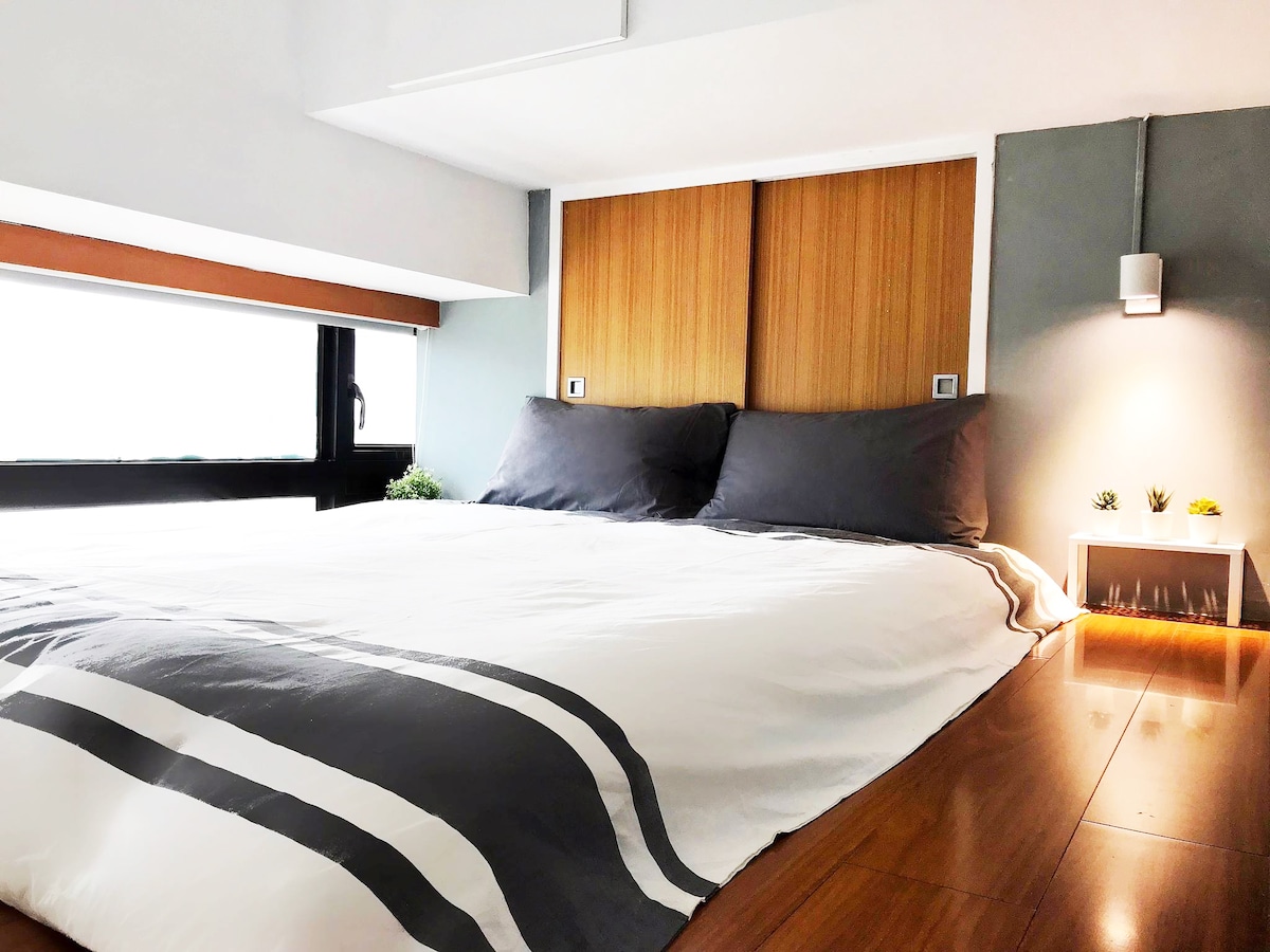 A comfortable sleeping area features a spacious bed dressed in a white duvet with subtle stripes. Light streams in through a large window, illuminating the wooden accents on the headboard and surrounding walls, creating a relaxed atmosphere.