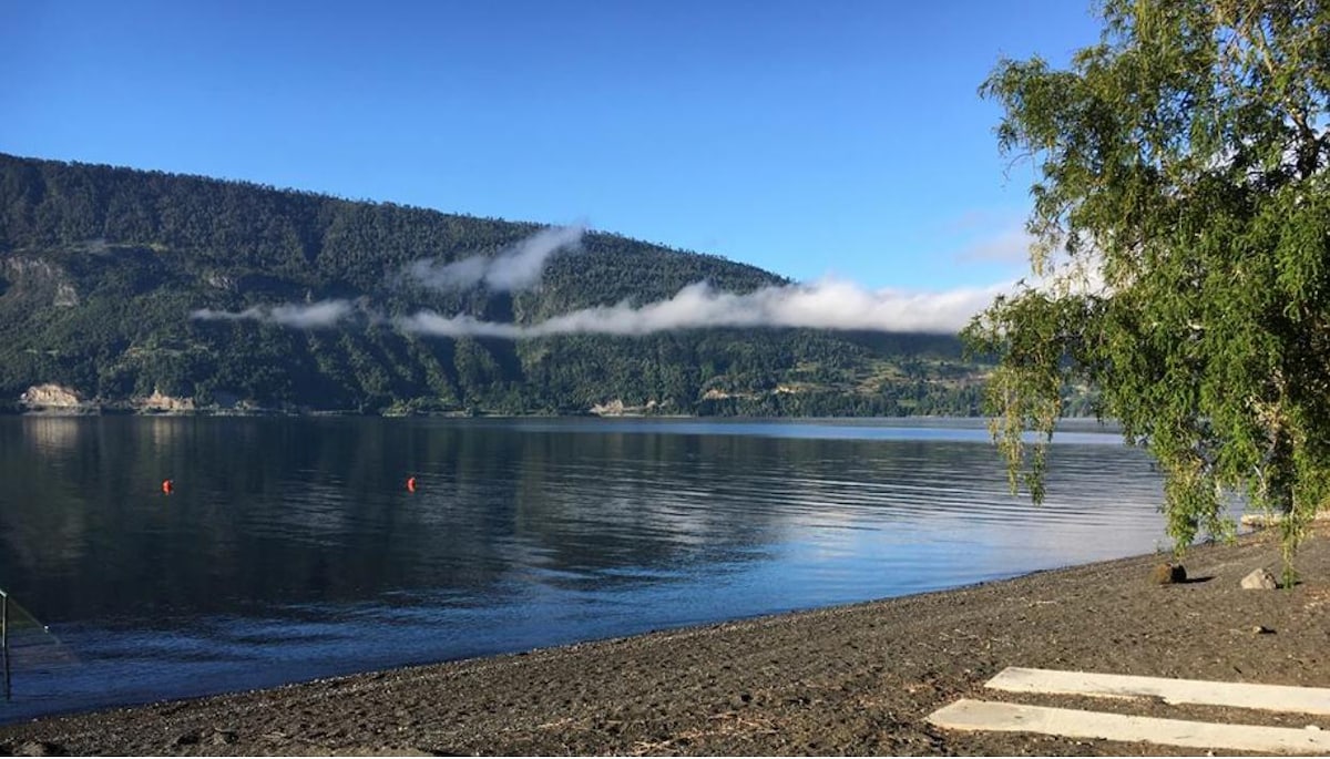 A serene lakeside view features calm waters reflecting the surrounding mountains and sky. The sandy shore is adorned with a few smooth stones and framed by greenery. A gentle mist rests on the hills in the background, creating a peaceful atmosphere.